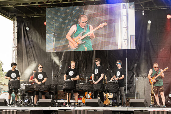 A rock band on an outdoor stage performing, with a large screen showing a guitarist in the background and players in black shirts in front of amplifiers.