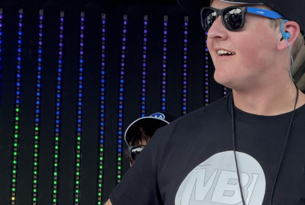 Screenshot 2026-08-21 at 34950 PM - MBI Arts Young man in a cap and sunglasses on stage, wearing headphones; shirt reads 'NBI Vibes' with a vertical LED backdrop behind him.