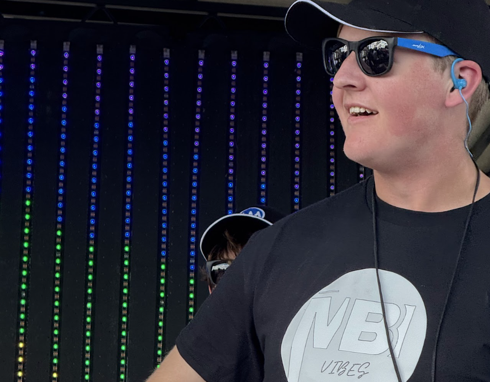 Young man in a cap and sunglasses on stage, wearing headphones; shirt reads 'NBI Vibes' with a vertical LED backdrop behind him.