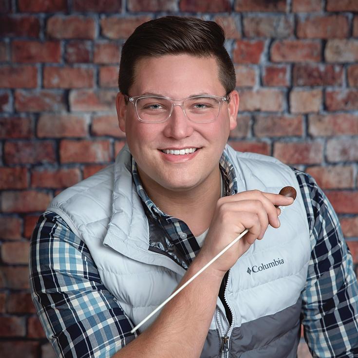 Smiling man with clear glasses, plaid shirt and light vest, holding a small cigar, against a brick wall in a casual portrait.