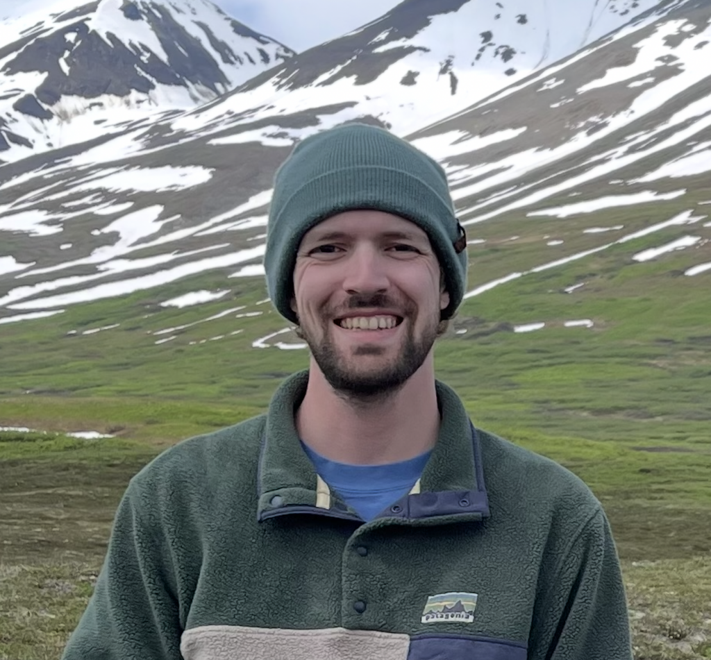 Smiling man in a green fleece and teal beanie with snowy mountains in the background.