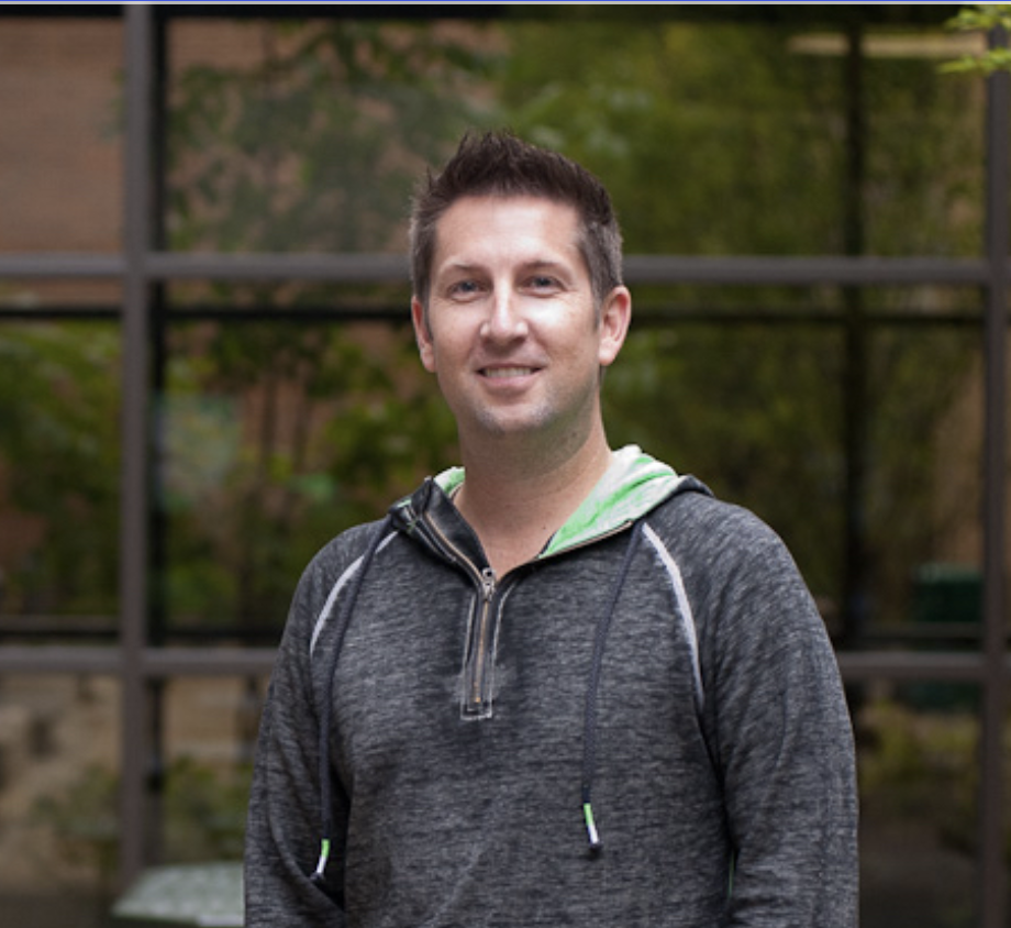 Portrait of a man in a gray zip-up hoodie with lime green lining, standing outdoors in front of a glass building.