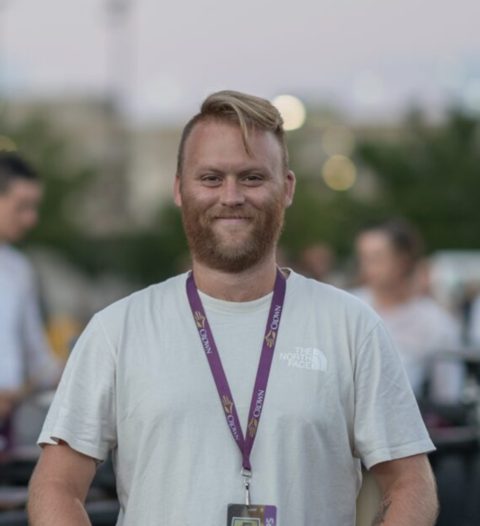 Smiling man wearing a light gray T-shirt and a purple lanyard with an ID badge at an outdoor event.
