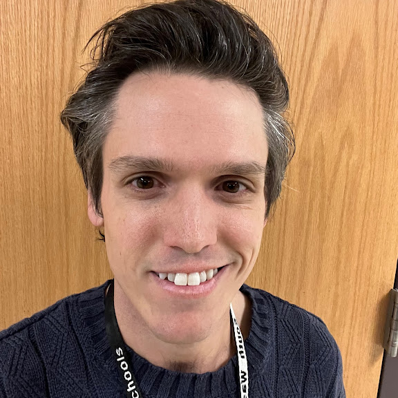Smiling man with dark hair wearing a navy knit sweater, standing in front of a wooden door.
