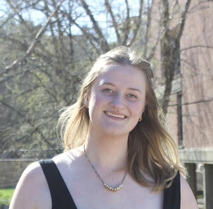 Smiling young woman with blonde hair outdoors, wearing a black dress and a necklace, in a sunny park setting.
