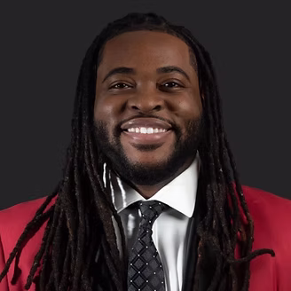 DeondreCarter - MBI Arts Portrait of a smiling man with long dreadlocks wearing a red blazer and a dark background behind him.