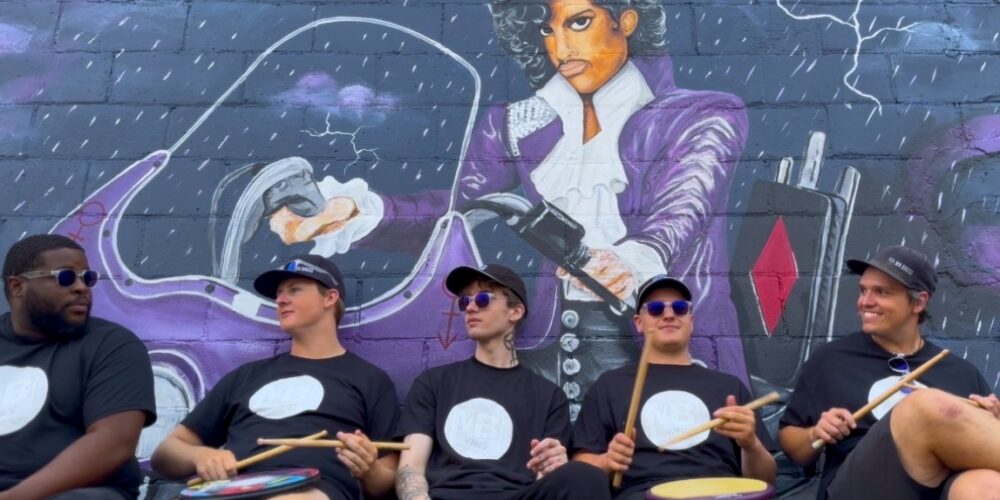 Five drummers in black shirts and sunglasses sit with sticks and handheld drums, posed in front of a purple mural depicting a musician in a suit.