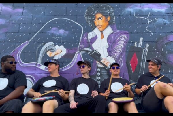 Five drummers in black shirts and sunglasses sit with sticks and handheld drums, posed in front of a purple mural depicting a musician in a suit.