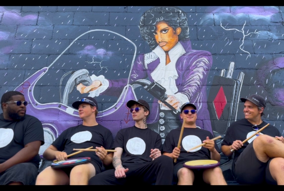 Five drummers in black shirts and sunglasses sit with sticks and handheld drums, posed in front of a purple mural depicting a musician in a suit.