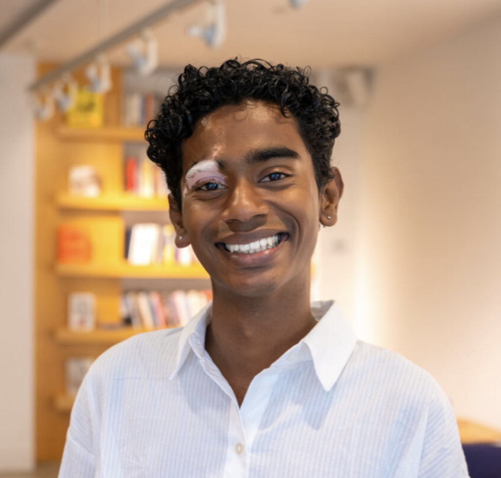 Smiling person with short curly hair in a light blue shirt, standing in a bright indoor space with bookshelves in the background; left eye has a white patch.
