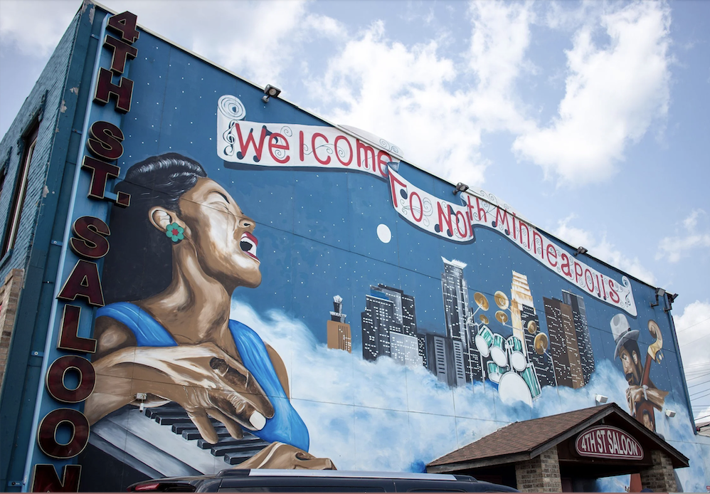 Screenshot 2026-08-21 at 125550 PM - MBI Arts Mural on a blue building: a woman singing at a piano with a city skyline and a 'Welcome to North Minneapolis' banner, plus a vertical '4th St Saloon' sign.