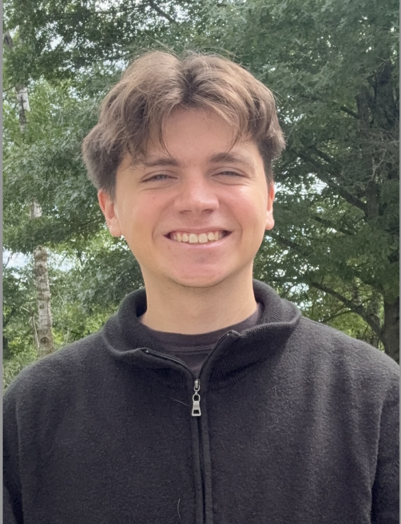 Young man smiling outdoors in a black zip-up fleece, with green trees in the background.