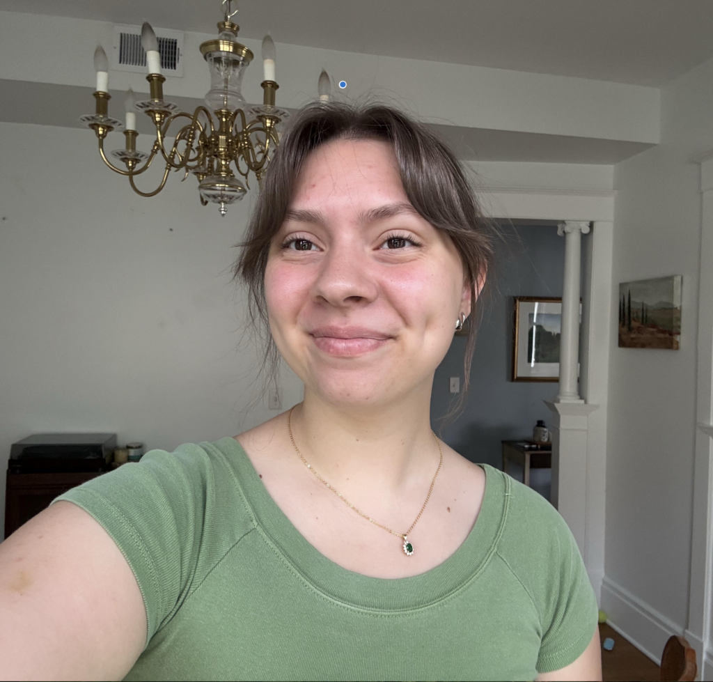 Smiling woman in a green top wearing a pendant necklace indoors, with a brass chandelier and framed art in the background.