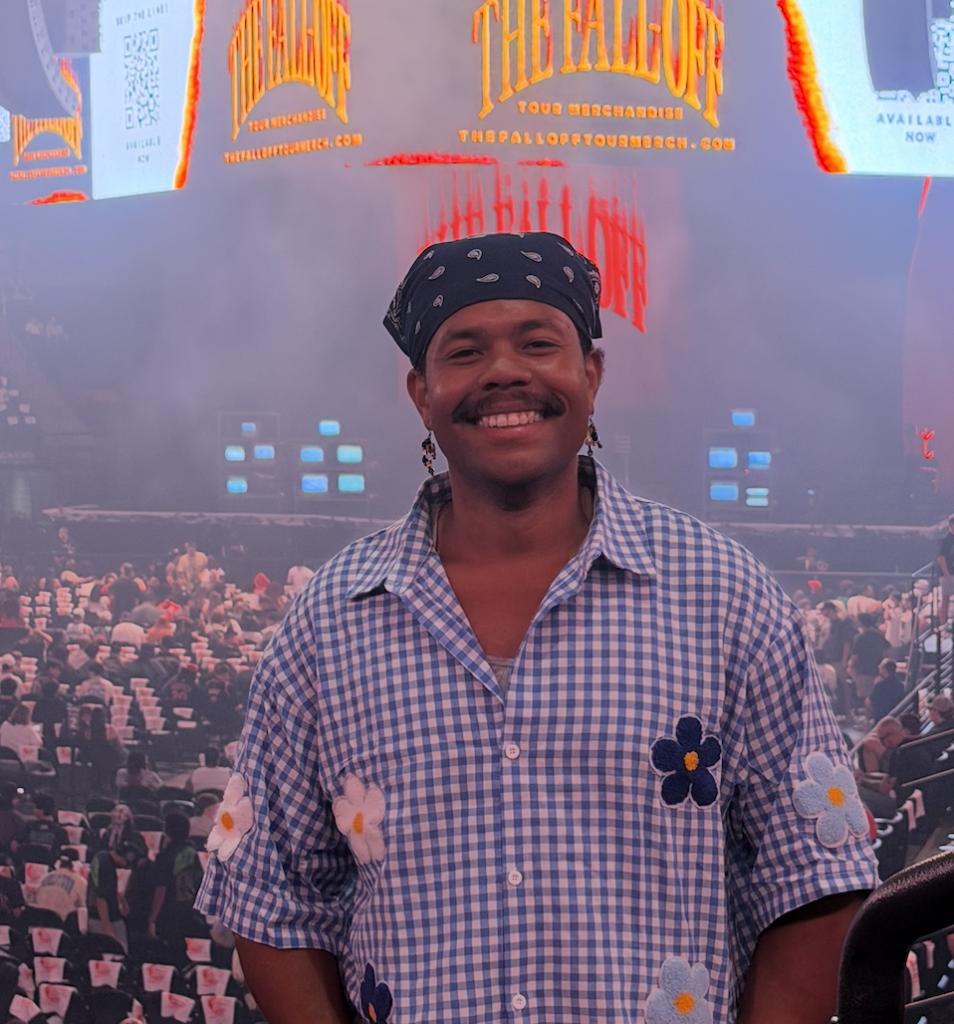 Smiling man in a blue gingham shirt with flower patches and a bandana, posing in front of a crowded concert venue with bright stage lights.