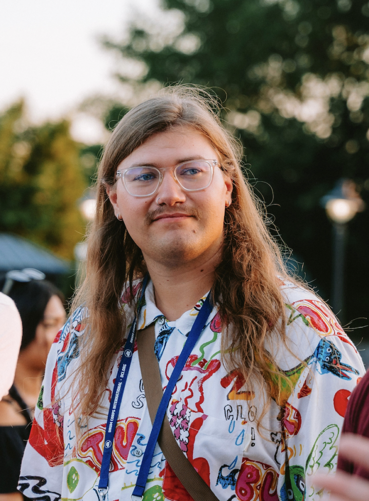 Person with long hair and clear glasses wearing a colorful printed shirt at an outdoor event.