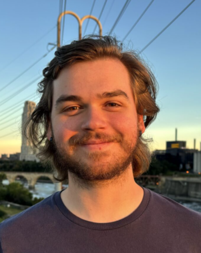 Young man with shoulder-length brown hair and beard smiling, sunset sky behind him with a city skyline and a bridge.
