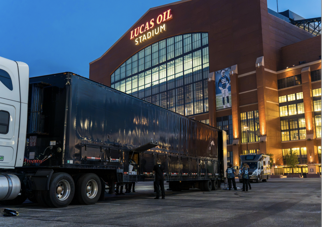 Night view of Lucas Oil Stadium with a large black semi-trailer parked in the foreground and workers nearby.