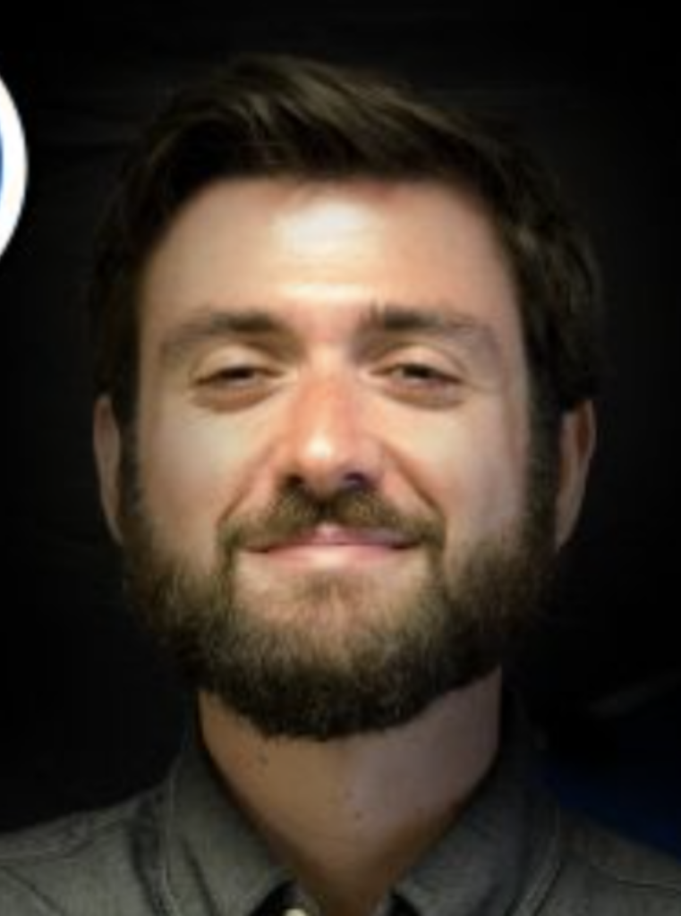 Smiling man with dark hair and a beard, wearing a gray shirt, facing the camera against a dark background.