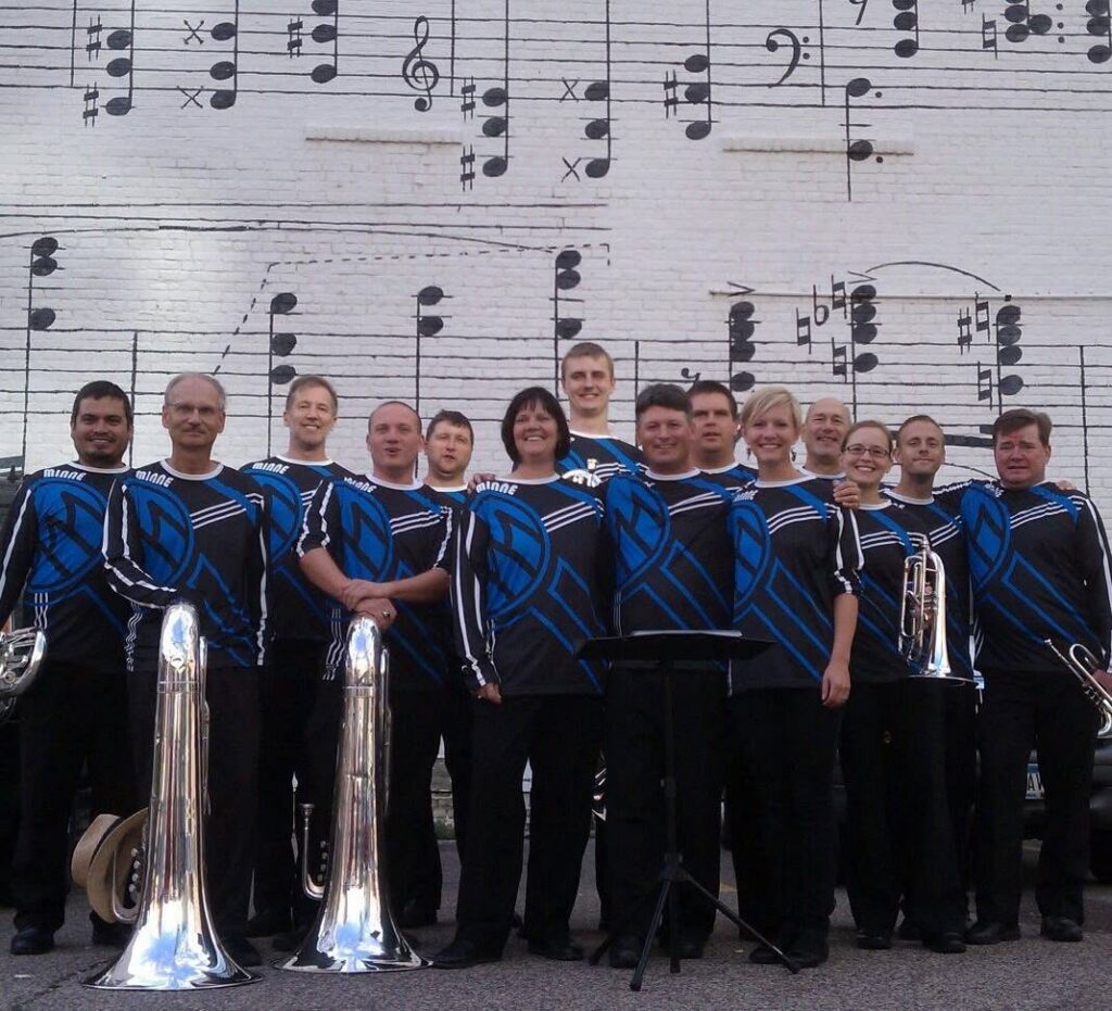 minne-brass-marquette-music - MBI Arts Brass ensemble posing in matching blue and black uniforms with tubas and trumpets in front of a large wall mural showing musical notation and staff lines.