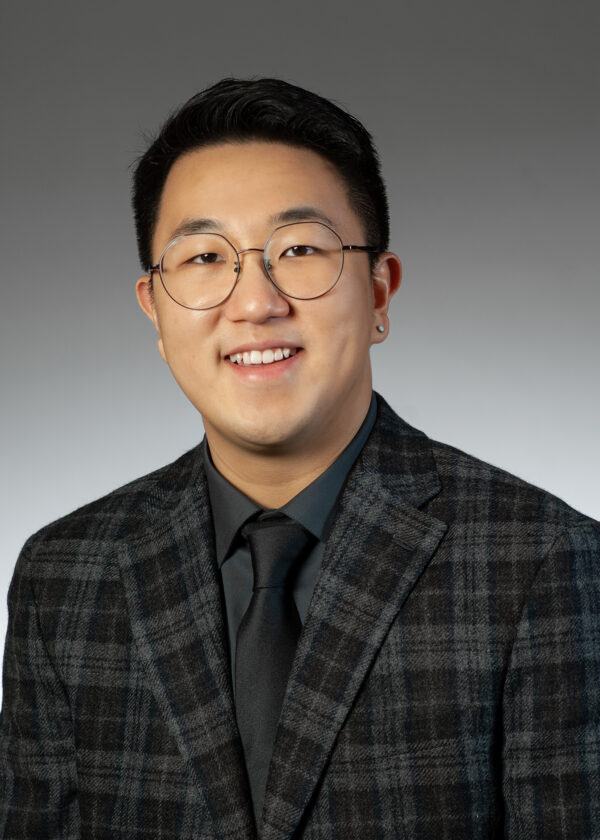 Portrait of a smiling man wearing round glasses and a dark plaid blazer.