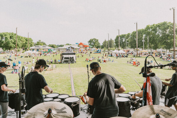 Back view of a percussion ensemble on an outdoor stage at a sunny park festival, with a crowd on the grass behind them.