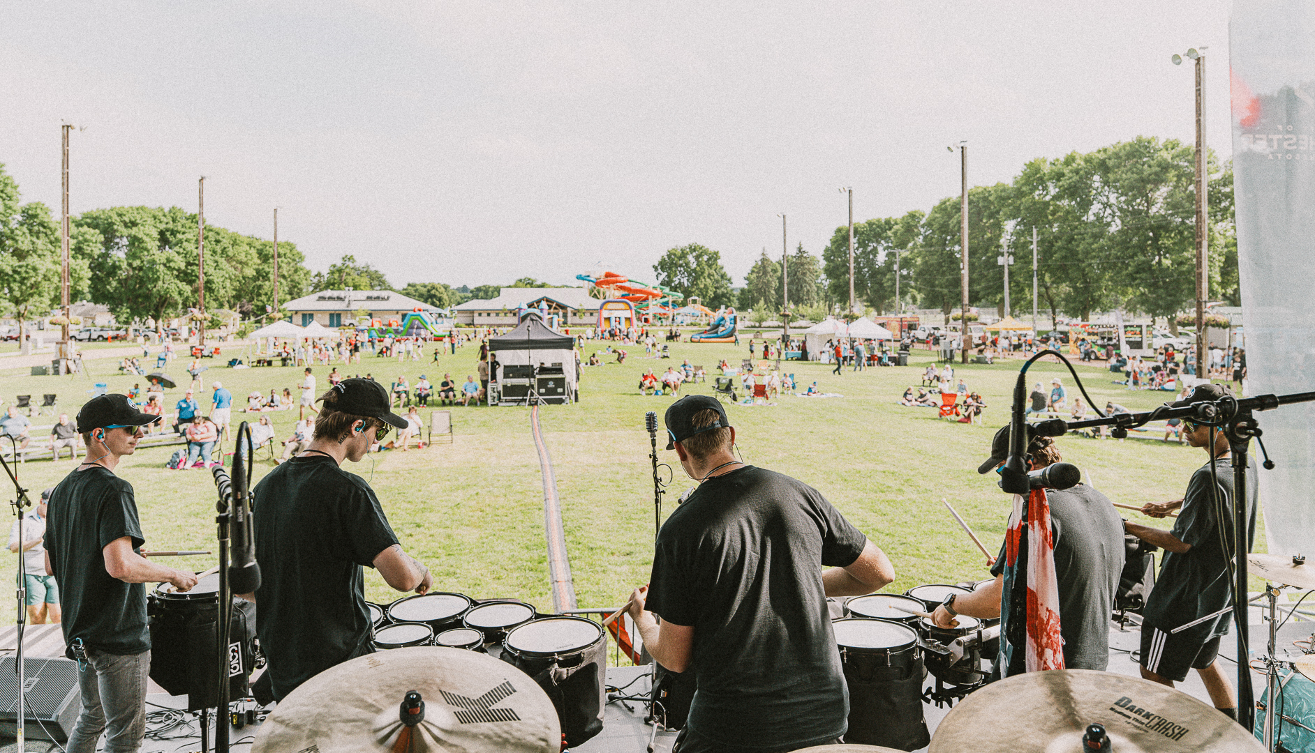Back view of a percussion ensemble on an outdoor stage at a sunny park festival, with a crowd on the grass behind them.
