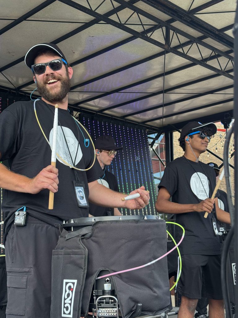 Bearded man in a cap and sunglasses on stage, holding drum sticks during a live performance with other musicians in the background.