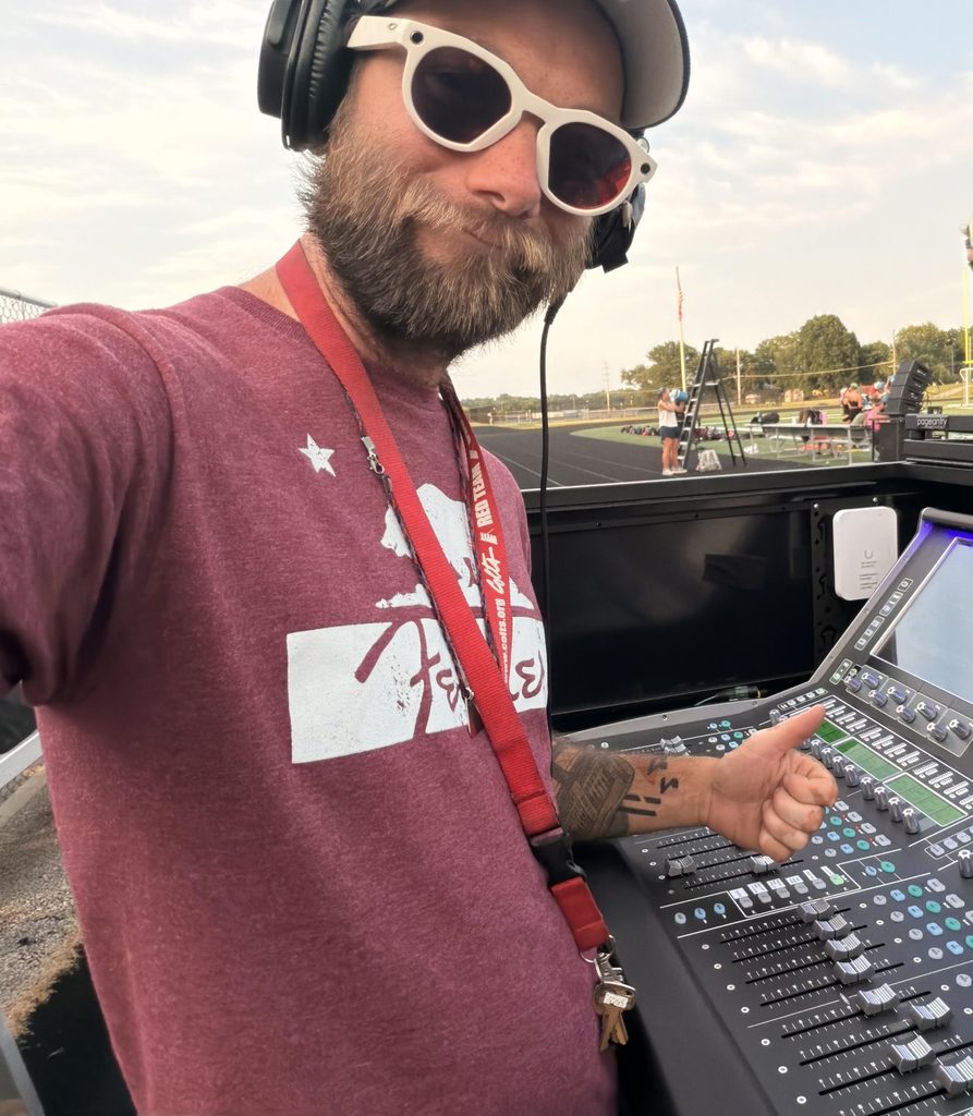 Josh Brulet Headshot - MBI Arts Man with a beard wearing white sunglasses and over-ear headphones operates a large outdoor sound mixing console. A red lanyard and keys hang from his neck as he adjusts controls in a stadium setting.