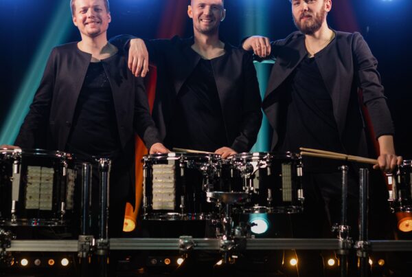 Three male drummers stand behind a row of drums on a lit concert stage, smiling at the camera in matching dark outfits.