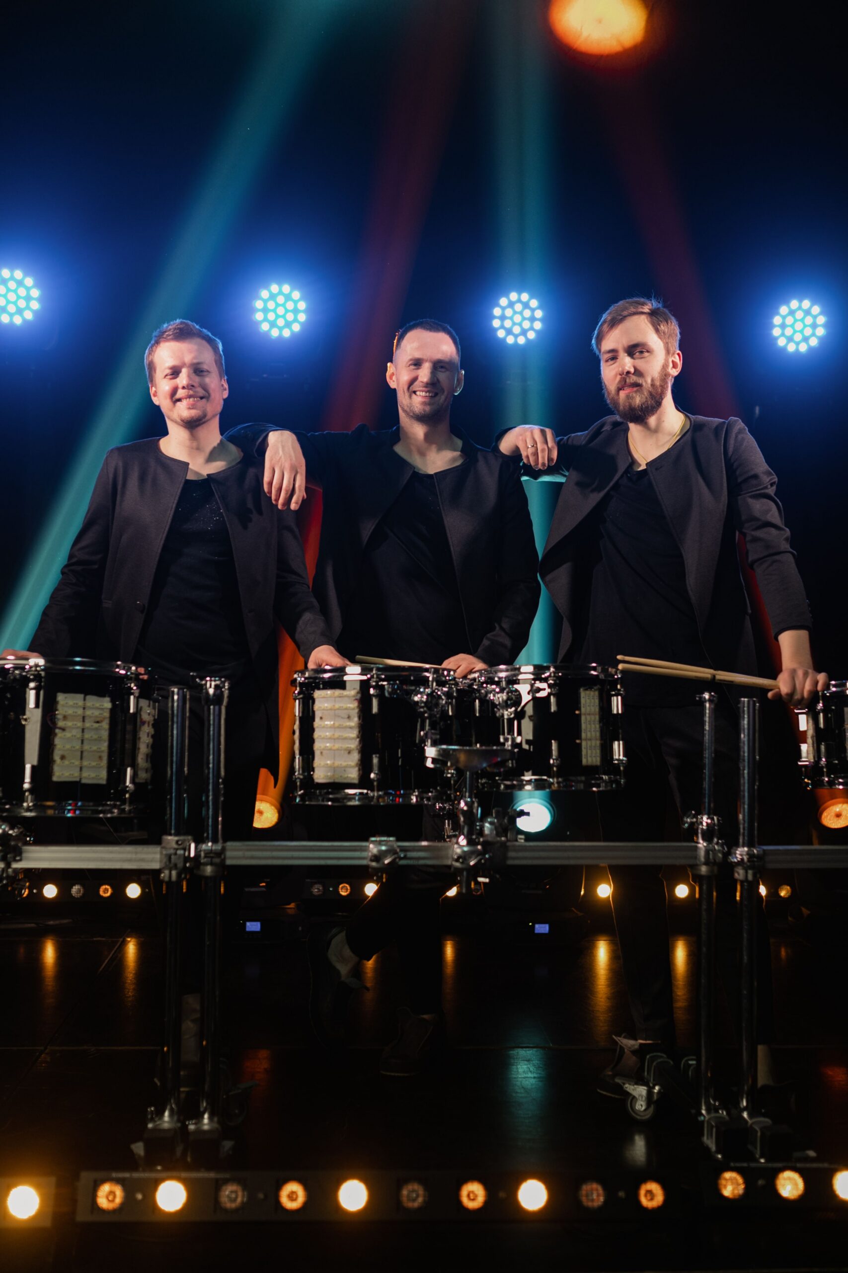 Three male drummers stand behind a row of drums on a lit concert stage, smiling at the camera in matching dark outfits.